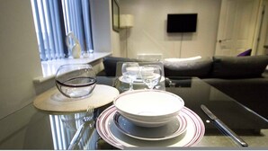 A dining area with a glass table, plates, glasses, and cutlery. There is a sofa and a TV in the background.