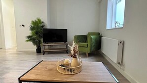 A modern living room with a flat-screen TV, a green armchair, a wooden coffee table with decorative items, and a potted plant.