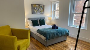 Une chambre à coucher avec un lit, un fauteuil jaune, un tableau, une fenêtre et une lampe.