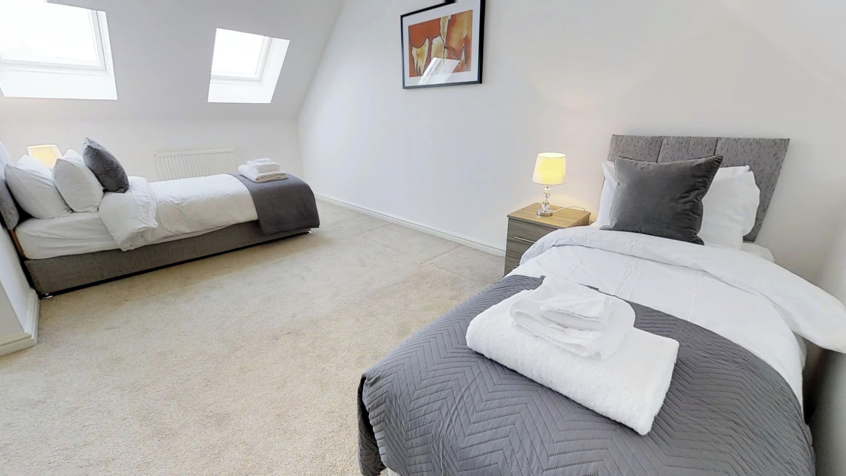 Une chambre avec deux lits, un radiateur, une table de chevet avec une lampe et un tableau au mur.