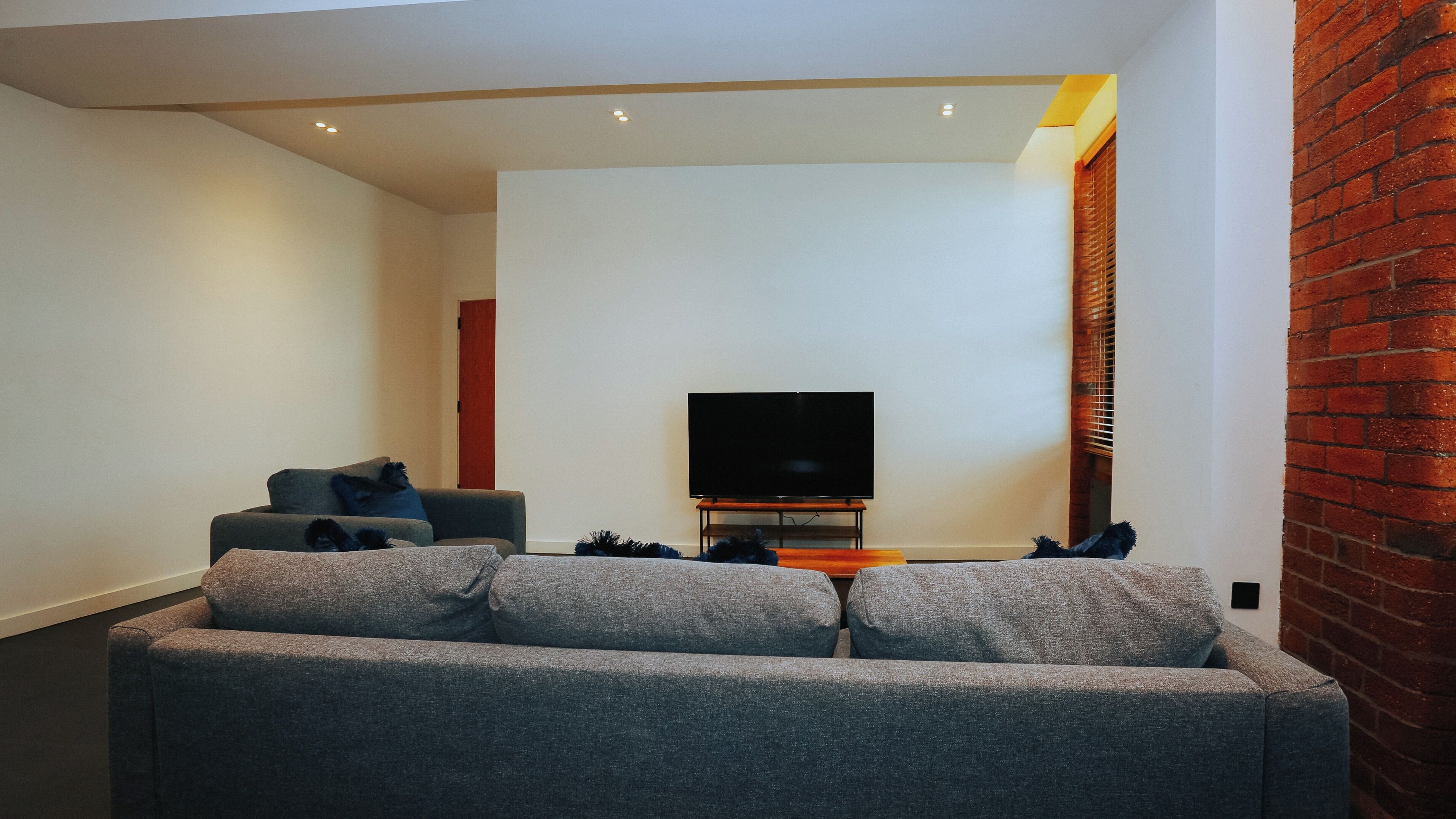 A modern living room with a grey sofa, a flat-screen TV on a stand, and a brick accent wall.