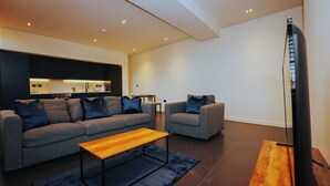 Een moderne woonkamer met een grijze bank, een kleinere fauteuil en een houten salontafel. Een flatscreen televisie is aan de muur bevestigd.