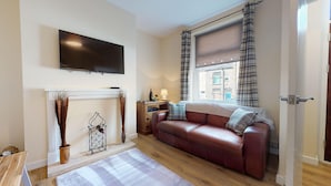 A living room with a fireplace, a sofa, a TV mounted on the wall, and a window with curtains. - Stayzo Westminster Stylish House Accommodation (Bradford)