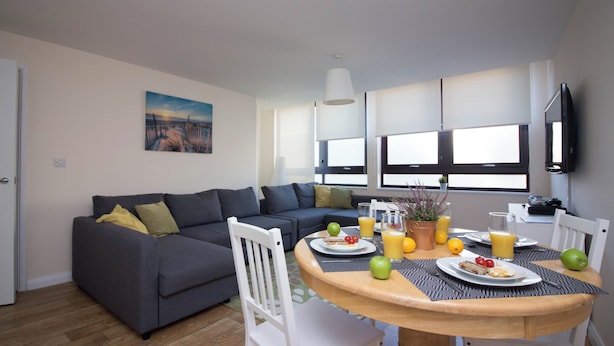 Una sala de estar moderna con un sofá gris, una mesa de comedor con alimentos para el desayuno y un ventanal con persianas.