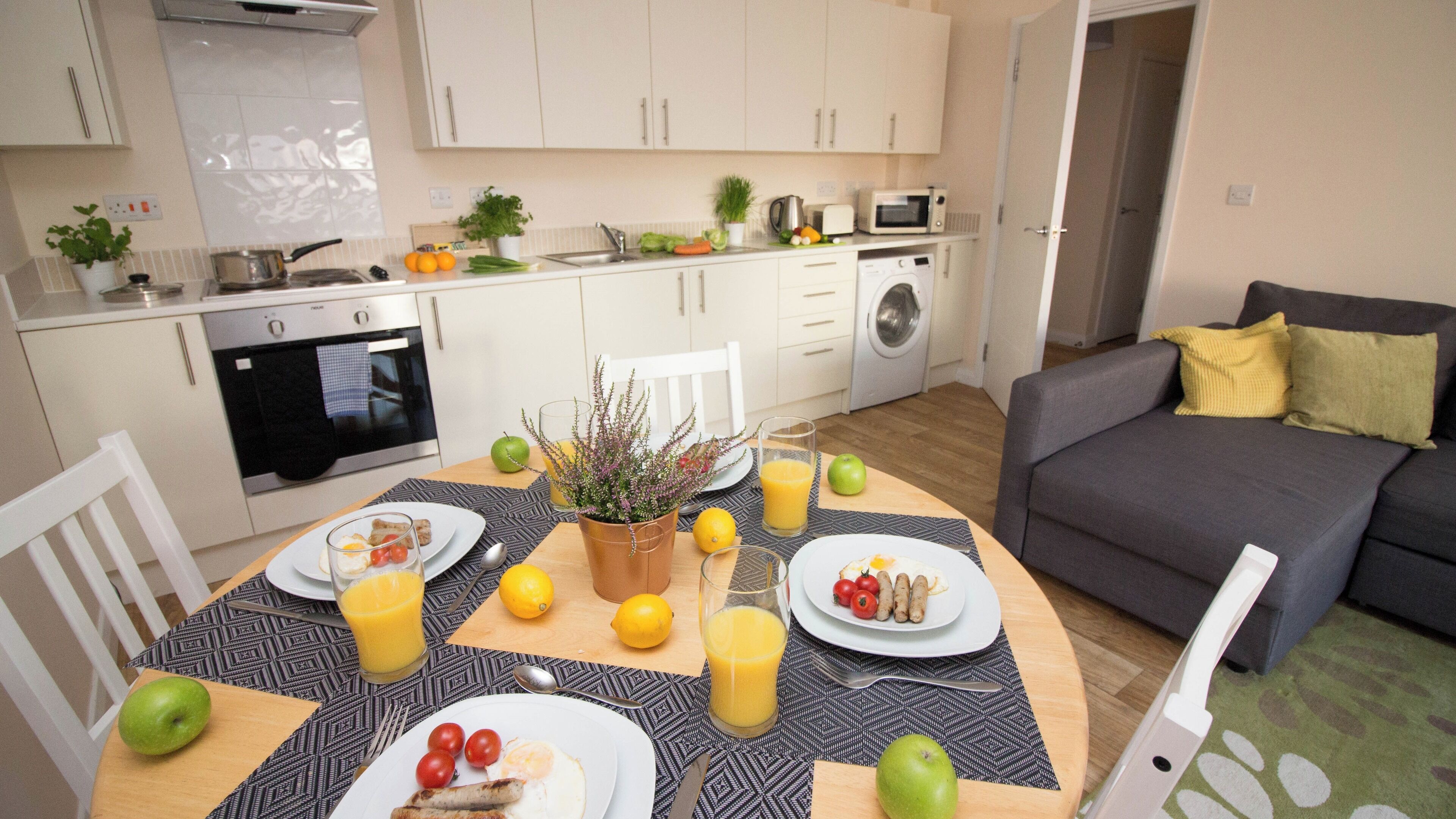 A modern kitchen with white cabinets, a dining table set with plates, glasses, and fruits, and a grey sofa in the living area.