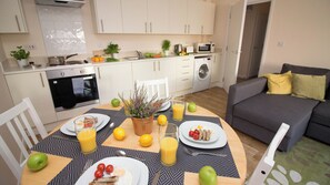 Una cucina moderna con mobili bianchi, un tavolo da pranzo apparecchiato con piatti, bicchieri e frutta, e un divano grigio nella zona soggiorno.
