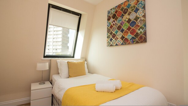 Une chambre à coucher bien rangée, avec un lit, une lampe de chevet, une fenêtre avec des stores et une décoration murale colorée.