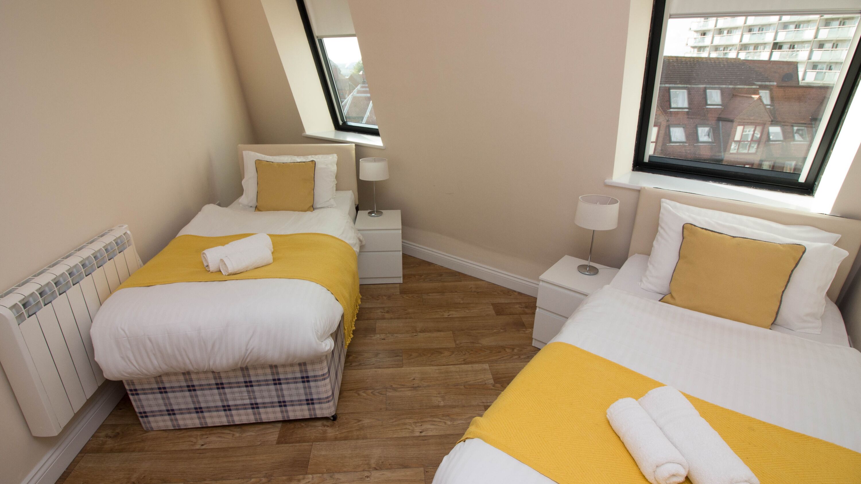Une chambre avec deux lits, chacun agrémenté de linge de lit blanc et de touches de couleur jaune, une table de chevet avec une lampe, et une vue sur les bâtiments par la fenêtre.
