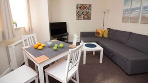 A living room with a grey sofa, white chairs, a small table with fruit, a TV, and artwork on the wall.