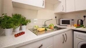 A kitchen with white cabinets, a stainless steel sink, a microwave, and various vegetables on the counter. - Stayzo Castle Point 18 Apartments - Premier Lodge (Southampton)