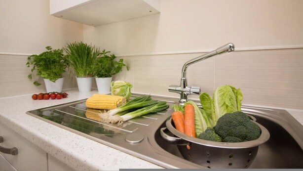 A kitchen with a sink, colander, and various vegetables like broccoli, carrots, and corn. - Stayzo Castle Point 18 Apartments - Premier Lodge (Southampton)