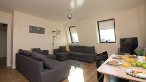 A modern living room with a sectional sofa, a dining table with breakfast items, and a large window providing natural light. - Stayzo Premiere Serviced Accommodation - 17 (Southampton)