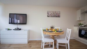 Une cuisine compacte avec un coin repas, un téléviseur à écran plat fixé au mur et une petite table à manger agrémentée de fruits et d’un vase.