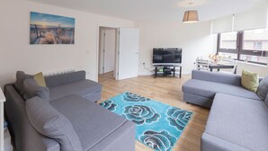 Un salon moderne avec deux canapés gris, un tapis à motifs floraux bleus, un téléviseur à écran plat et un tableau représentant un coucher de soleil.