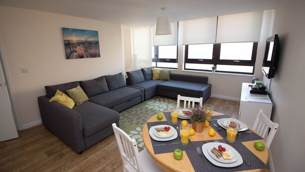 Un salon moderne avec un canapé d’angle gris, une table à manger dressée pour le petit-déjeuner et un tableau accroché au mur.