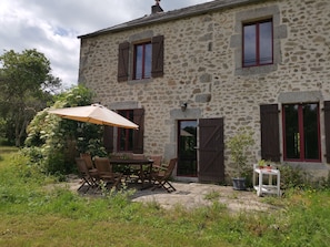 A stone building with a patio area featuring a table and chairs, a potted plant, and a small cart. - Anjali's House in Morvan (Saint-Martin-du-Puy)