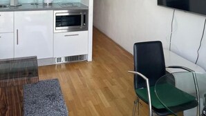 Une cuisine moderne avec des meubles blancs, un four à micro-ondes et une machine à café. On y trouve une table en verre avec des chaises, ainsi qu’un téléviseur à écran plat fixé au mur.