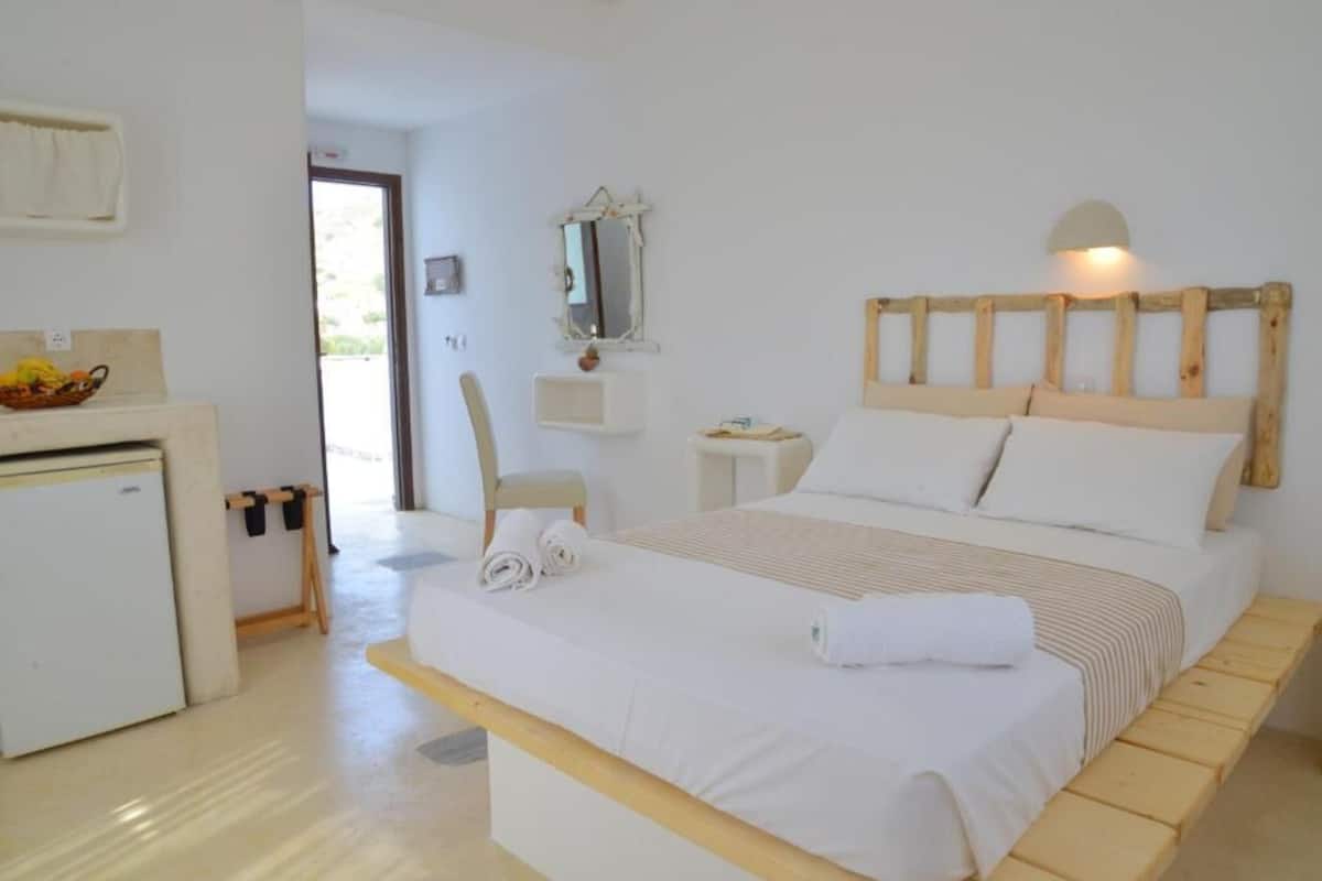 A bedroom with a wooden headboard, a bed with white linens, a small bedside table, a mirror, and a white desk with a basket of fruit.