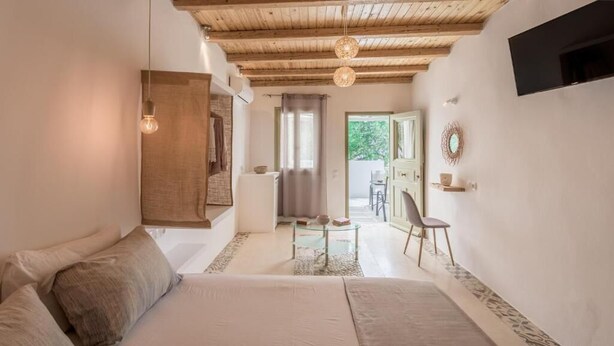 A bedroom with a bed, a small table with a chair, a wooden ceiling, and a view of the outside. - Fantasy Rooms  (Milos)