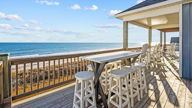 A wooden deck with a table and stools overlooking the ocean.
