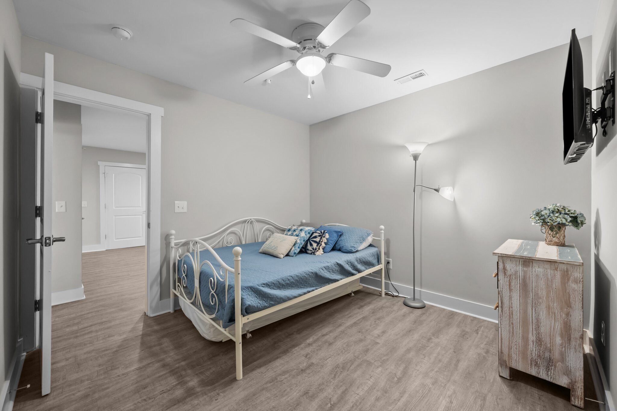 Une chambre à coucher comprenant un lit, un ventilateur de plafond, une lampe de chevet, une petite armoire en bois et une porte donnant sur une autre pièce.