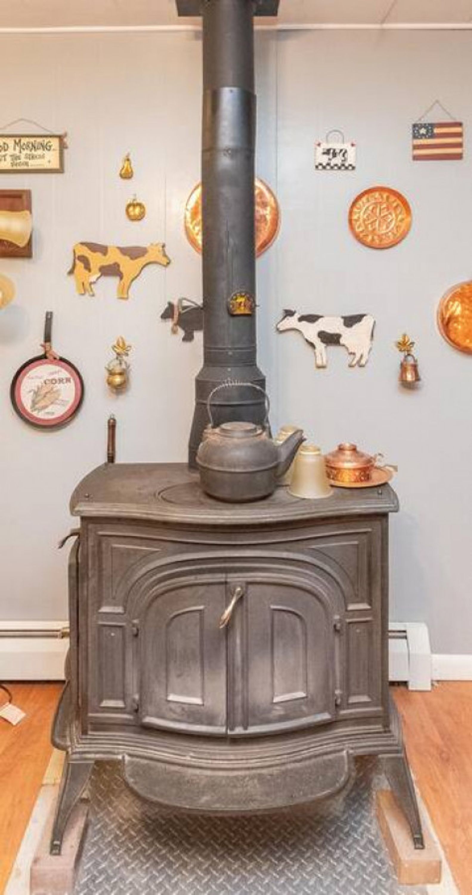 Una estufa de leña tradicional con chimenea negra, una olla metálica encima y un recorte decorativo de vaca en la pared.