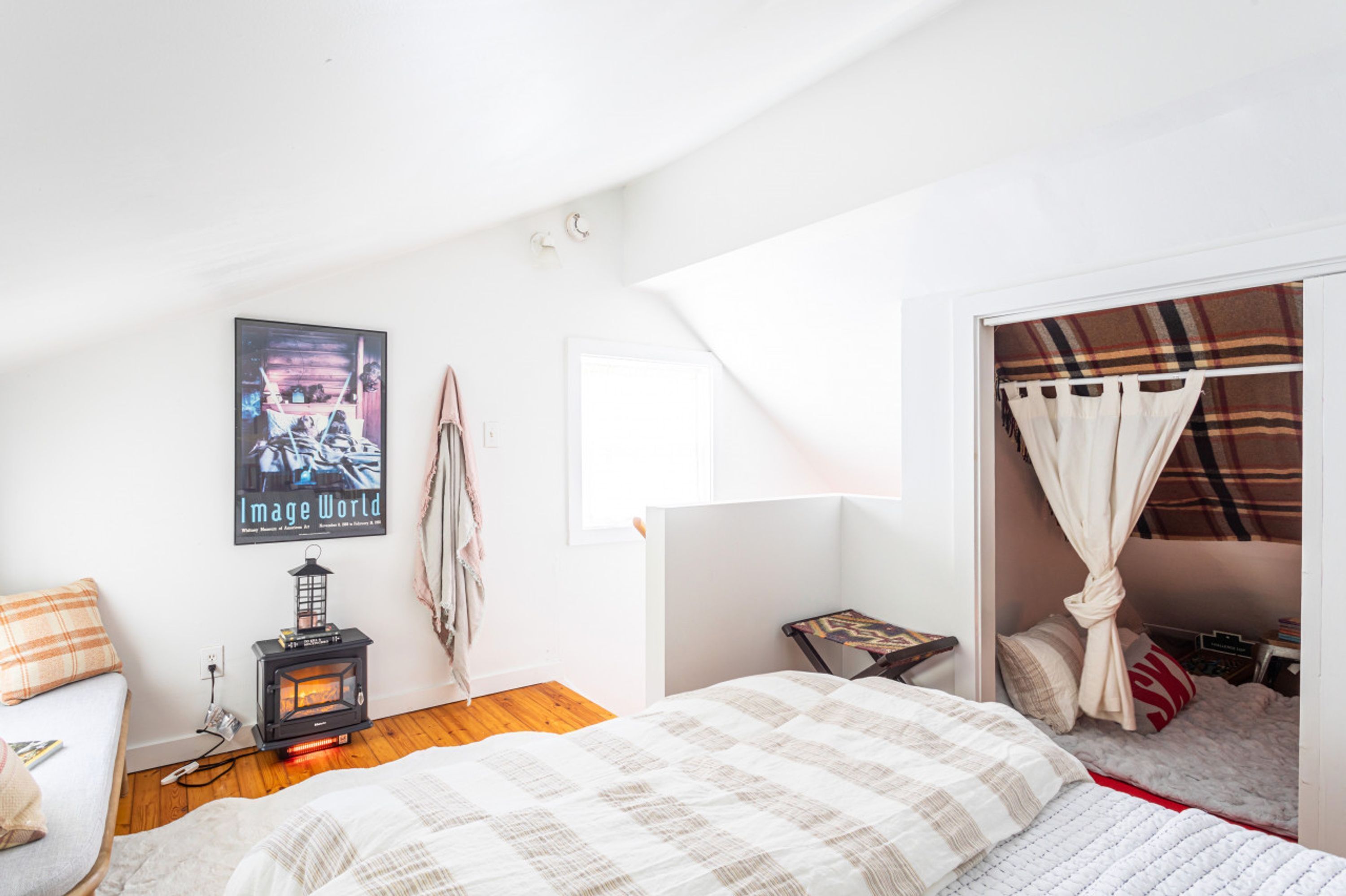 Un dormitorio con cama, una mesita, un calentador, una ventana con cortinas y un cartel en la pared.
