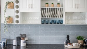 Une cuisine avec des meubles blancs, un dosseret en carrelage gris et divers ustensiles de cuisine.