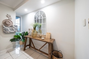 A hallway with a wooden console table, a large arched window, and decorative items including a hat, plants, and a welcome sign. - Kim's On Kara (Airlie Beach)