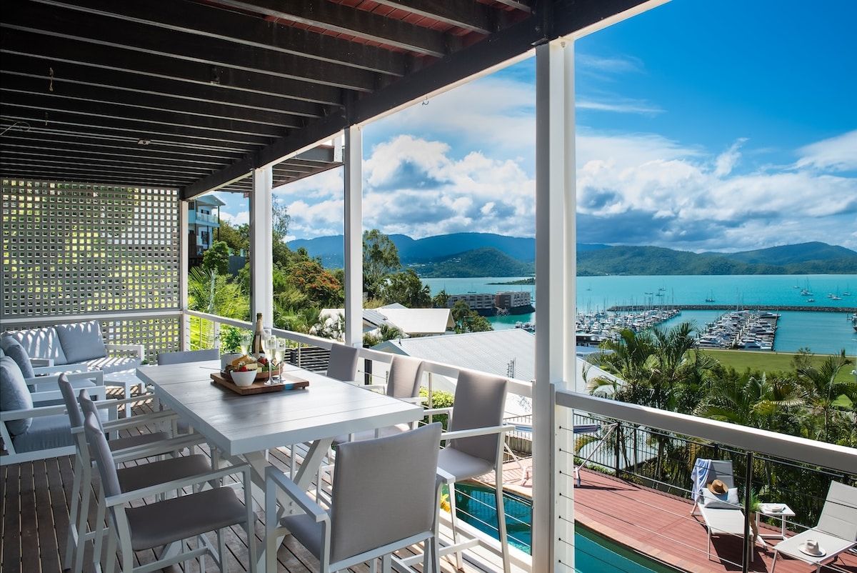 A balcony with a table and chairs, overlooking a marina and a body of water with mountains in the background.