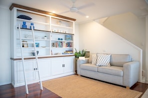 A living room with a white bookshelf, a grey sofa, a wooden floor, and a ceiling fan. - Kim's On Kara (Airlie Beach)
