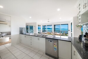 A modern kitchen with a view of the sea, white cabinetry, and a built-in dishwasher. - Kim's On Kara (Airlie Beach)