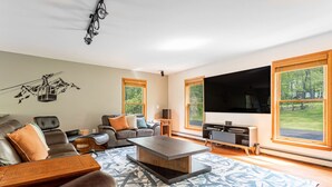 Ein modernes Wohnzimmer mit einem großen Flachbildfernseher, Ledersofas, einem Couchtisch und großen Fenstern mit Blick ins Freie.