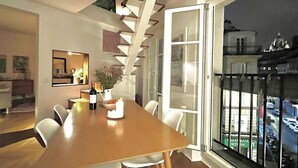 Uma sala de jantar com uma mesa de madeira comprida, cadeiras brancas, uma garrafa de vinho e um vaso com flores. Há uma escada que leva a um andar superior com corrimão de vidro.