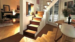 Um interior moderno com uma escada de madeira que leva a um segundo andar, uma área de jantar com mesa e cadeiras, e uma lareira ao fundo.