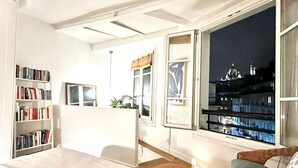 Quarto moderno com uma cama grande, uma estante, uma janela com vista para luzes da cidade e uma mesinha com uma vela.