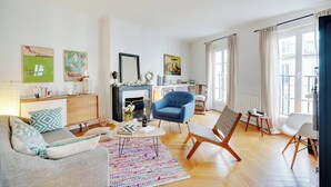 Uma sala de estar moderna com sofá, poltronas, mesa de centro e lareira. Há quadros nas paredes e uma janela com cortinas.