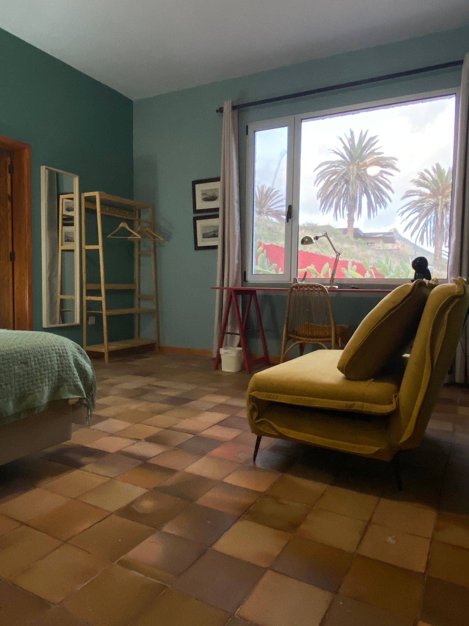 A room with a bed, a yellow armchair, a window with a view of palm trees, and a ladder against the wall.
