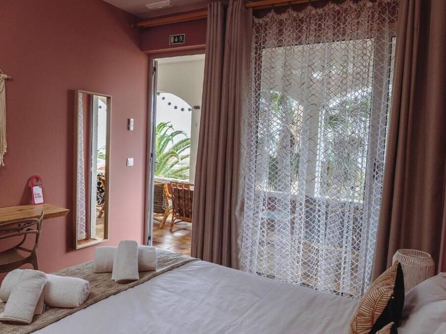 A hotel room with a bed, a desk, and a large window with lace curtains.