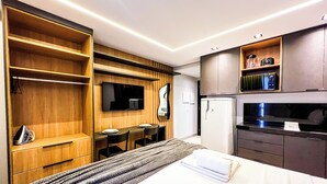 Moderni hotellihuone, jossa on sänky, seinälle asennettu televisio, pieni ruokapöytä tuoleineen, jääkaappi ja sisäänrakennetut puuhyllyt.