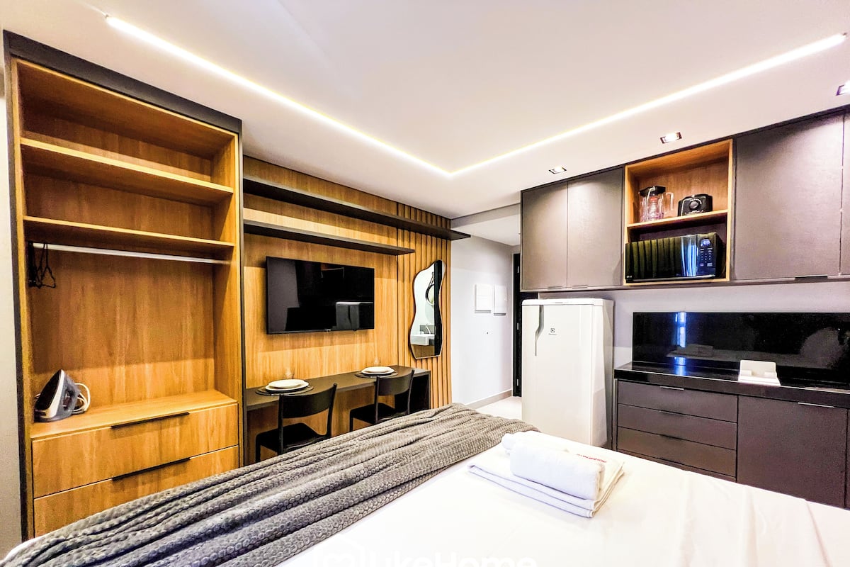 Une chambre d’hôtel moderne équipée d’un lit, d’un téléviseur fixé au mur, d’une petite table à manger avec des chaises, d’un réfrigérateur et d ’étagères en bois intégrées.