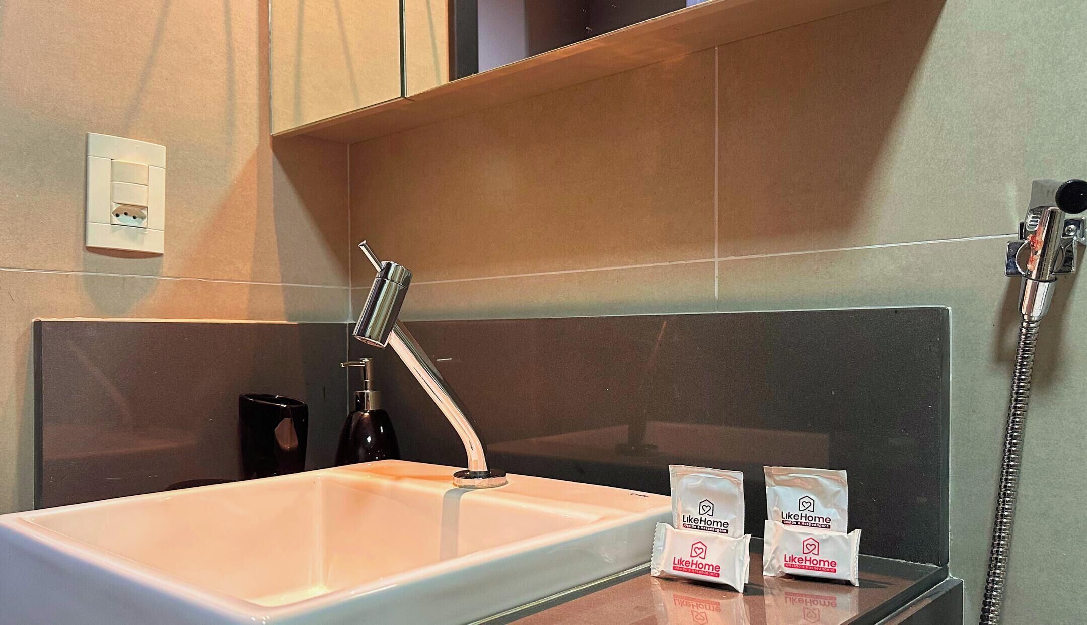 A bathroom with a white sink, a handheld showerhead, a soap dispenser, and two packages labeled 'LikeHome' on the counter.
