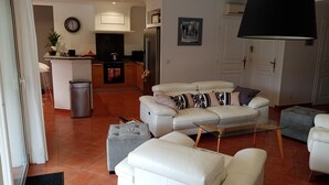 Un salon moderne avec un canapé, un pouf et un coin repas, la cuisine étant visible au fond.