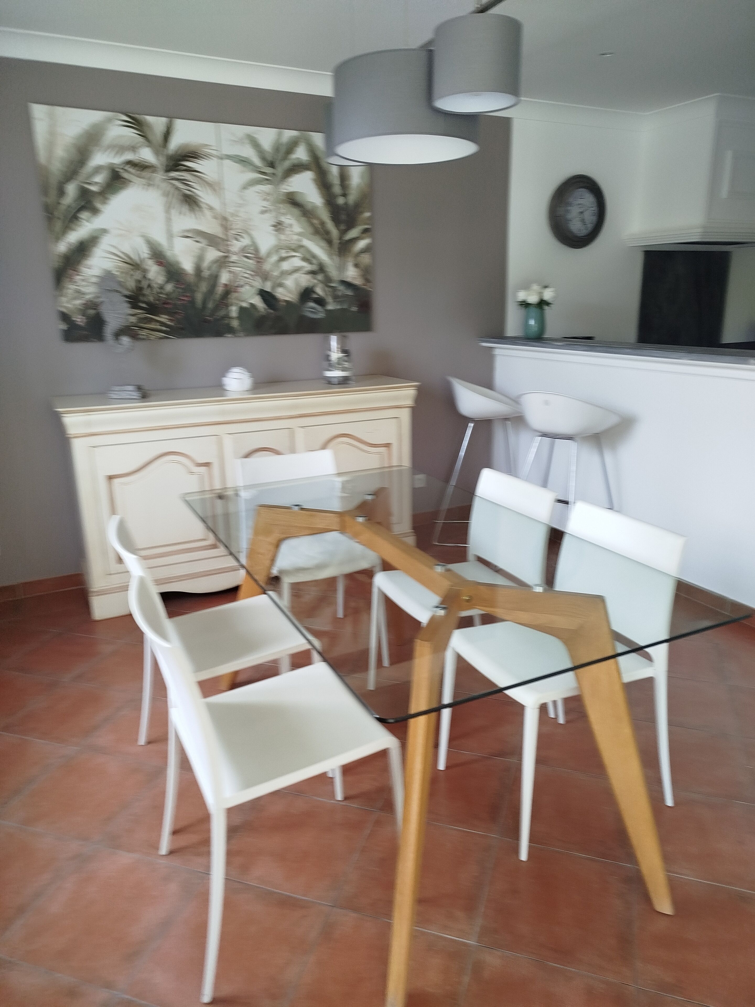 A dining area with a glass table and white chairs, a cream-colored cabinet, and a painting on the wall.