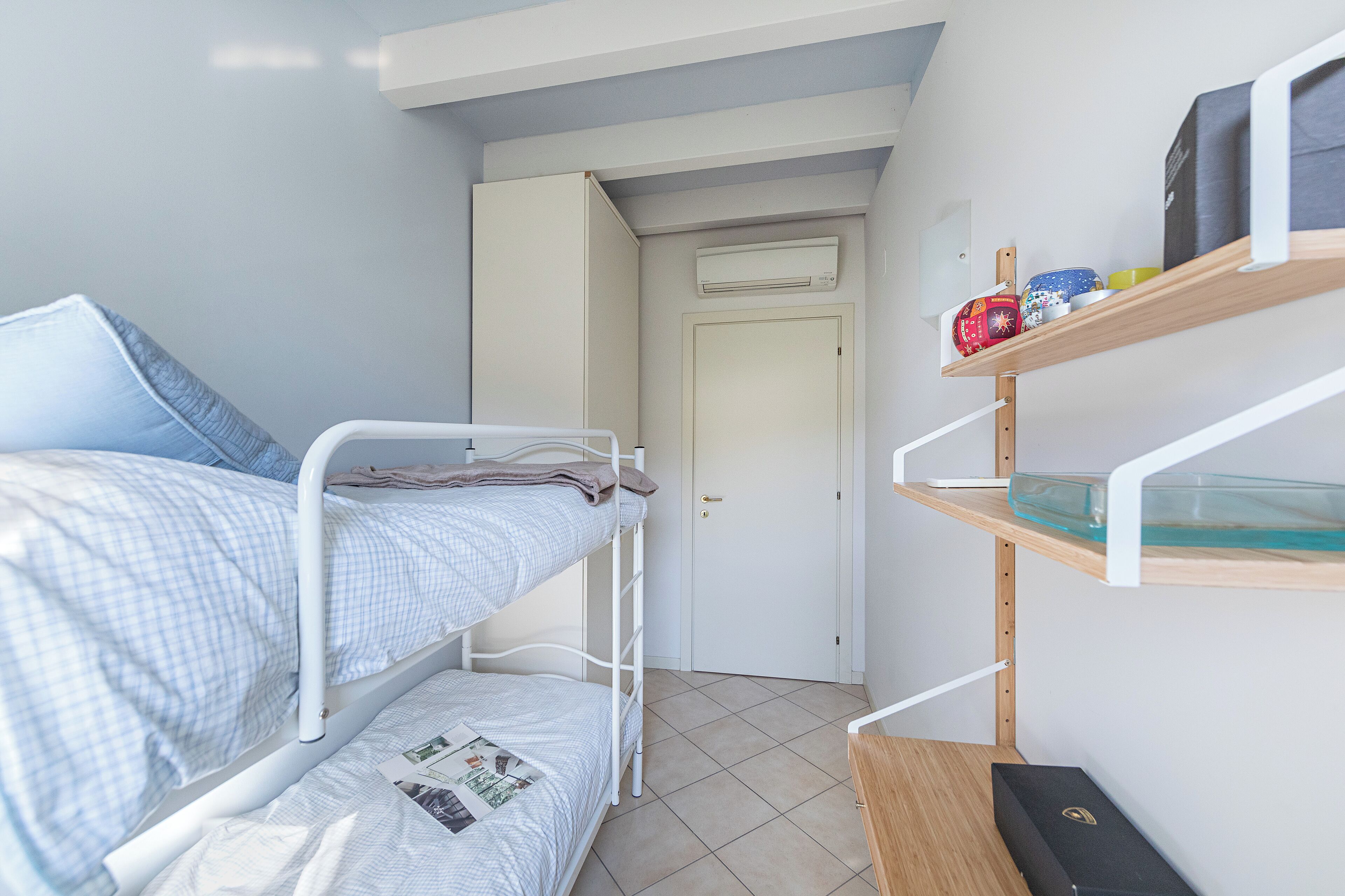 A small, well-lit room with bunk beds, a white door, and a shelf with various items.