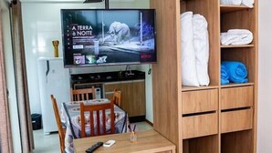 A hotel room with a bed, a wooden wardrobe, a TV on the wall, a desk with a chair, and a dining area with a table and chairs. - Residencial Canto dos Passaros (Florianópolis)