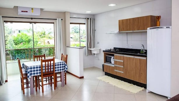 A modern kitchen with wooden cabinets, a white refrigerator, and a dining area with a checkered tablecloth. - Residencial Canto dos Passaros (Florianópolis)