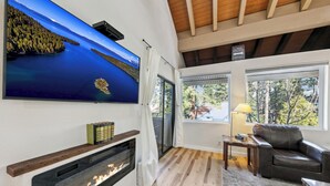 A modern living room with a fireplace, a large TV mounted on the wall, and a view of the outdoors through large windows. - Lakeland Village #309 Luxury Remodel 1BR+Loft (South Lake Tahoe)