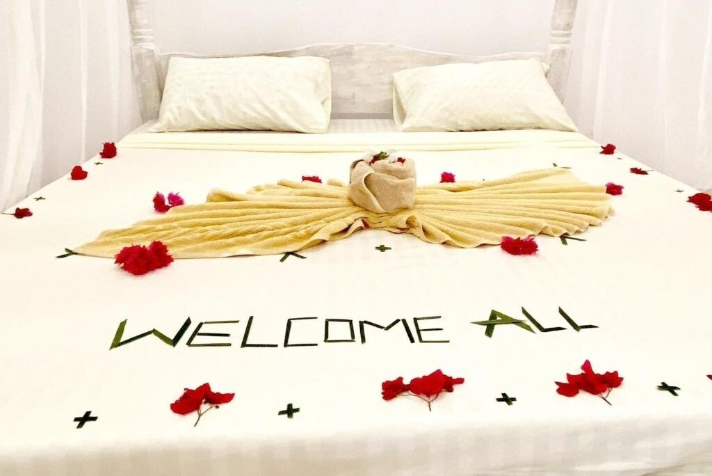A bed with a white bedspread, red flowers, and a towel arrangement spelling "WELCOME ALL".