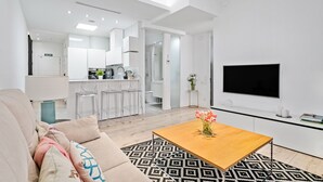 Ein modernes Wohnzimmer mit einer Couch, einem Couchtisch mit Blumen, einem Fernseher und einer Küchenzeile im Hintergrund.
